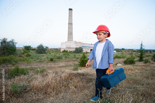 child labor concept of small kid in orange helmet with toolbox going along heavy industry plant factory with high pipe with copy space
