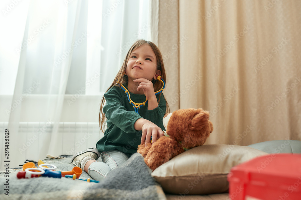 A small girl playing doctor with a toy stethoscope having a teddybear as a patient deciding what to do while sitting on a sofa in a spacy room A small girl playing doctor with a toy stethoscope having a teddybear as a patient deciding what to do while sitting on a sofa in a spacy room