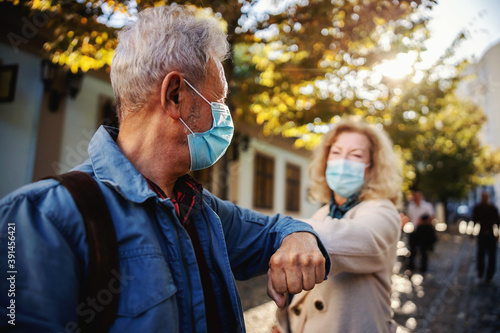 Senior friends with protective masks on standing outdoors and greet with their elbows. Although it's corona outbreak, we have friends.
