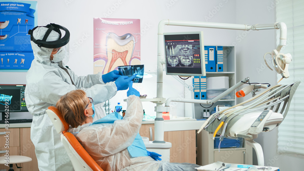Fototapeta premium Dentist with face shield reviewing panoramic mouth x-ray image to patient during global pandemic. Assistant and doctor talking with senior woman wearing suit, coverall, protection suit, mask, gloves