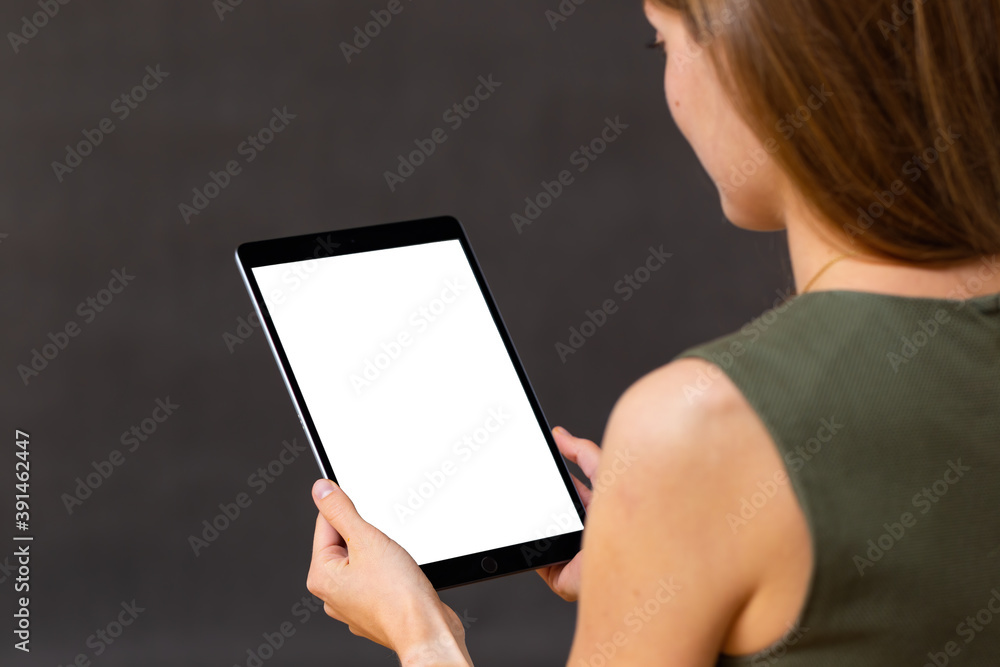 Interested woman holding tablet with white screen in portrait orientation in her hands on dark background. Look over shoulder from close-up of focused caucasian female using black mobile device.