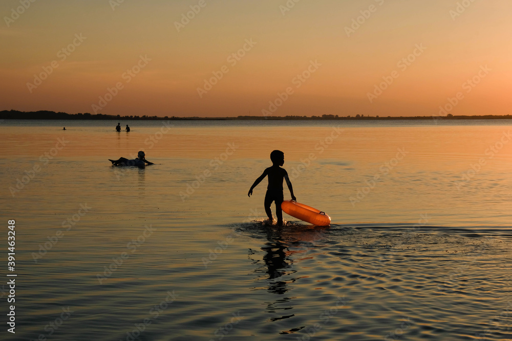 Children's silhouette on a sunset background. A child with an orange inflatable circle in shallow water. Vacation concept. Copy space.