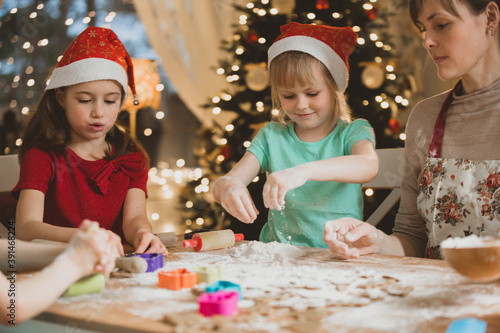 Mother and little kids in red hats cooking gingerbread cookies and playing. Beautiful living room with lights and Christmas tree  table with lantern. Happy family celebrating holiday together. 