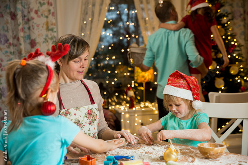 Mother and kids in red hats cooking gingerbread cookies and decorating glaze. Father with child decorating Christmas tree. Beautiful living room with lights. Happy family celebrating holiday together.