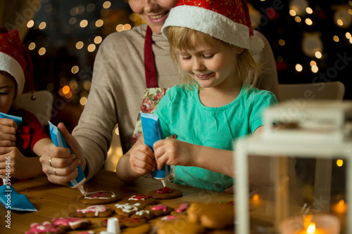 Mother and little kids in red hats cooking gingerbread cookies and decorating with glaze. Beautiful living room with lights and Christmas tree. Happy family celebrating holiday together.