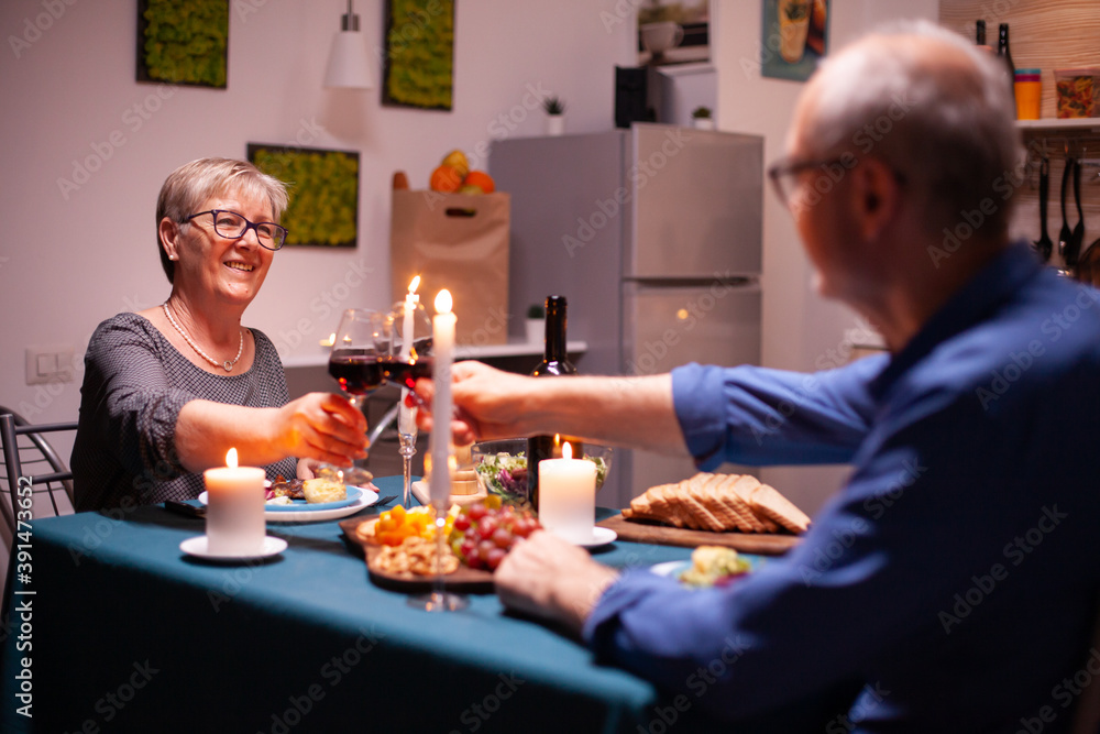 Happy pensioner couple toasting with red wine glasses in kitchen. Senior couple sitting at the table in kitchen, talking, enjoying the meal, celebrating their anniversary in the dining room. Happy pensioner couple toasting with red wine glasses in kitchen. Senior couple sitting at the table in kitchen, talking, enjoying the meal, celebrating their anniversary in the dining room.
