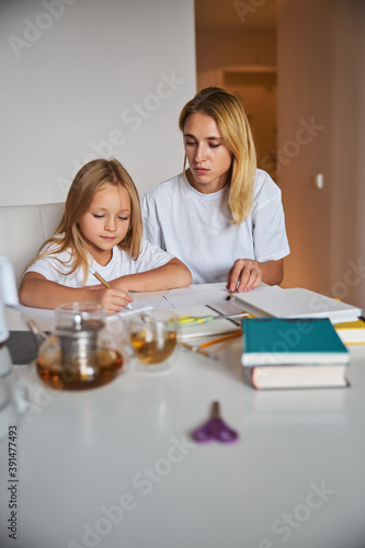 Thinking little girl writing something in the exercises book with mom in room inside