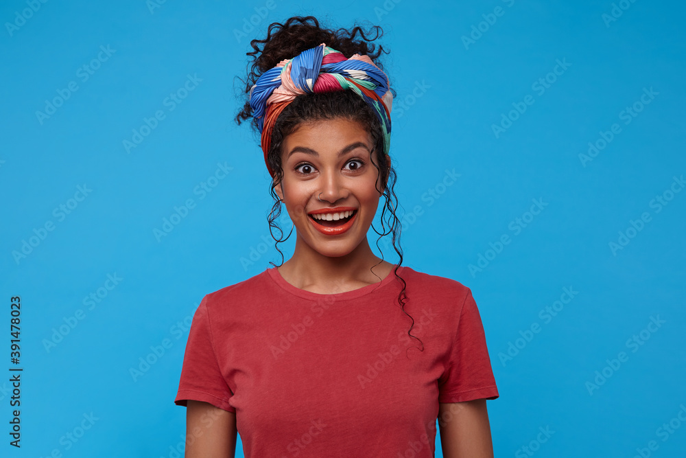 Agitated young attractive brunette curly woman with casual hairstyle raising surprisedly eyebrows while looking excitedly at camera, isolated over blue background