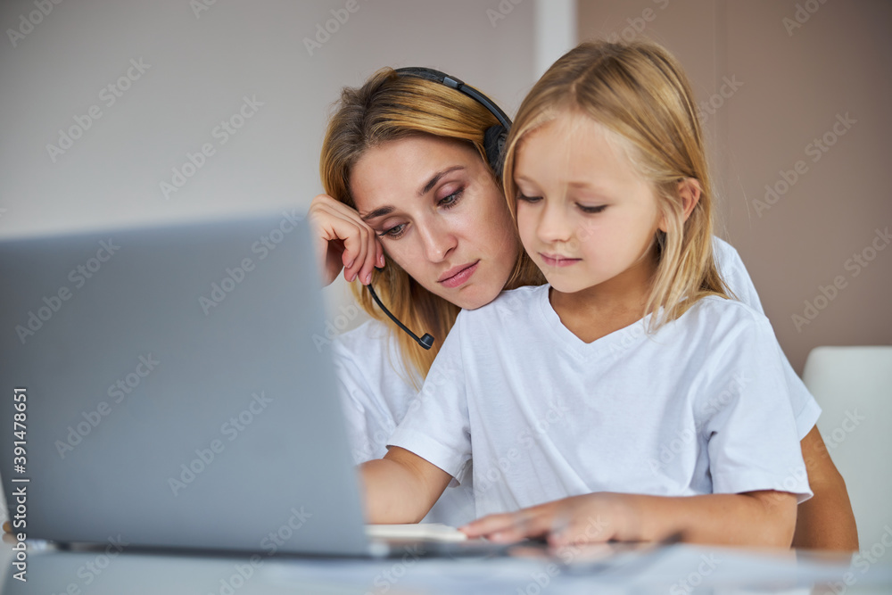 Sad mother in headphone spending time with her daughter while sitting at the desk in room inside Sad mother in headphone spending time with her daughter while sitting at the desk in room inside