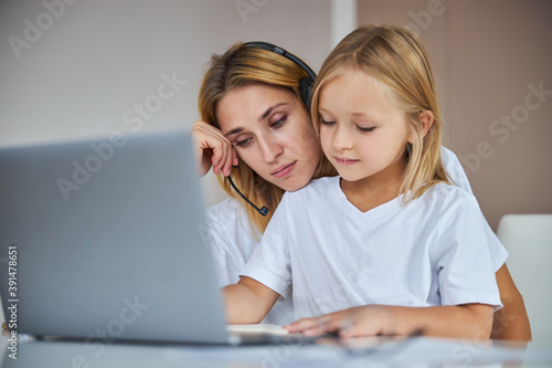 Sad mother in headphone spending time with her daughter while sitting at the desk in room inside