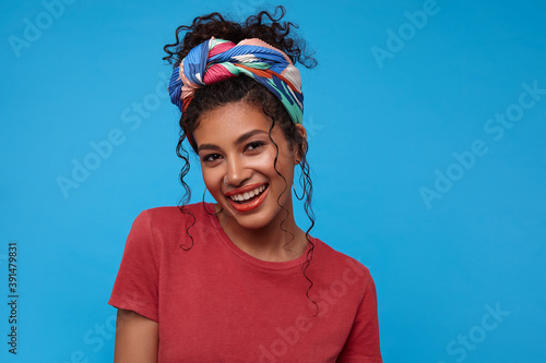 Beautiful young pretty dark haired curly woman with casual hairstyle looking gladly at camera and smiling broadly while standing over blue background