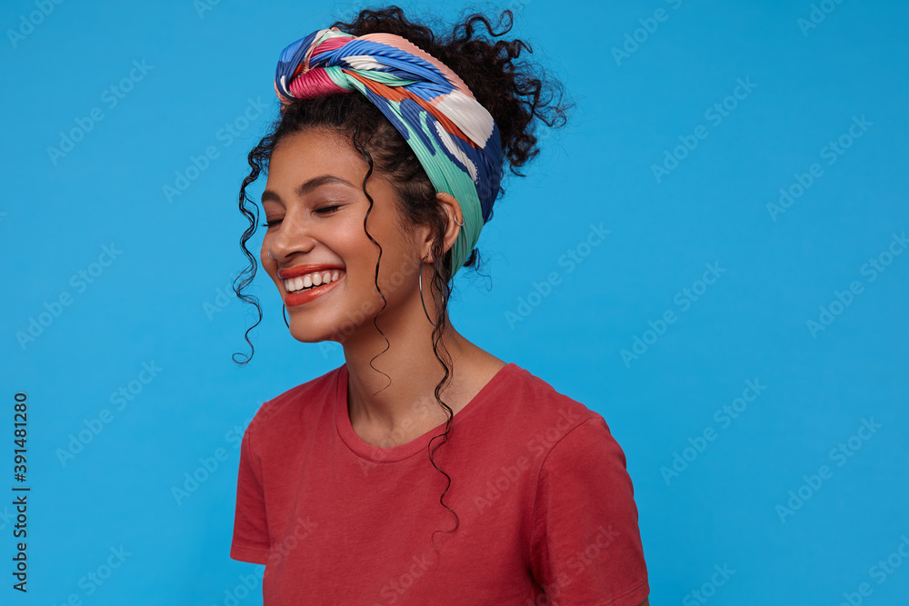 Good looking happy young brunette curly woman with multi-colored headband being in nice mood and miling gladly with closed eyes while standing over blue background Good looking happy young brunette curly woman with multi-colored headband being in nice mood and miling gladly with closed eyes while standing over blue background