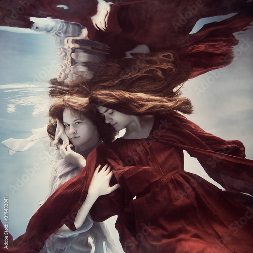 Two girls underwater in blue and red dresses with developing long hair seem to fly in weightlessness and interact with each other