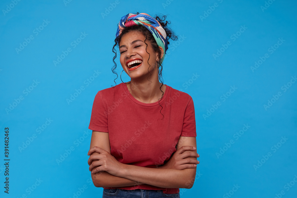 Happy young lovely dark haired woman with gathered hair throwing back her head while laughing happily, standing over blue background with folded hands