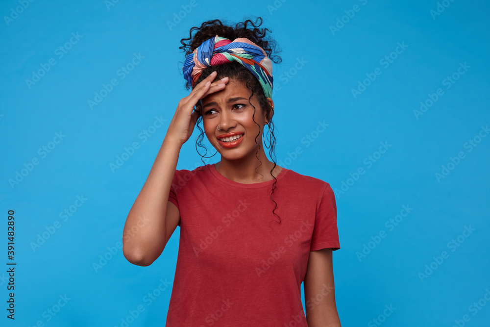 Indoor photo of young pretty dark haired curly lady with festive makeup frowning her face and keeping raised hand on forehead while posing over blue background