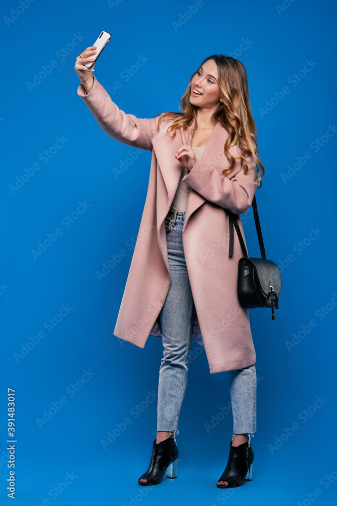 .Young beautiful blonde girl in a raincoat on a blue background with mobile phone doing selfie .Young beautiful blonde girl in a raincoat on a blue background with mobile phone doing selfie
