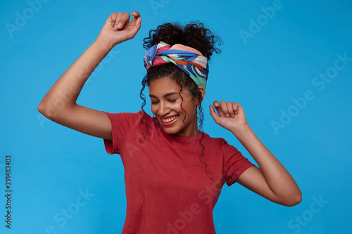 Good looking young pretty brunette curly woman with gathered hair smiling cheerfully with closed eyes while dancing with raised hands, posing over blue background