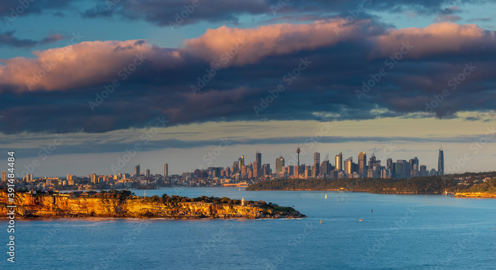 Panoramic view of the Sydney harbour during the sunrise. Clouds and rocks are catching the first lights of the rising sun, city skyscrapers in the background.