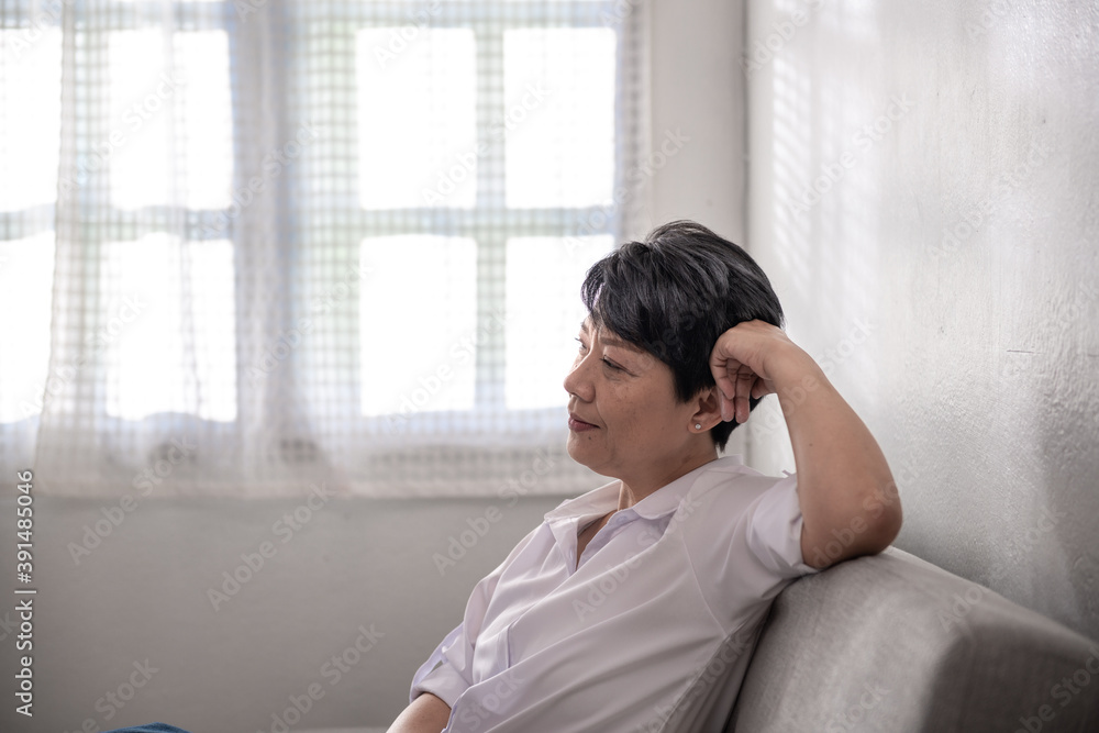 Happy Middle-aged gray-haired Asian woman wearing a white shirt poses at home on a sunny morning, a single retired woman sitting on the sofa in the living room. Positive single senior retired Concept Happy Middle-aged gray-haired Asian woman wearing a white shirt poses at home on a sunny morning, a single retired woman sitting on the sofa in the living room. Positive single senior retired Concept