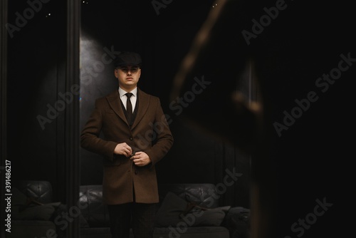 A man posing in the image of an English retro gangster of the 1920s dressed in a coat, suit and flat cap in Peaky blinders style.