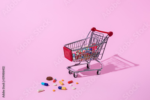 Creative medical layout made with pharmaceutical medicine pills, tablets, capsules in shopping trolley on pastel pink background. Minimal modern pharmacy or health care concept with trend shadows.