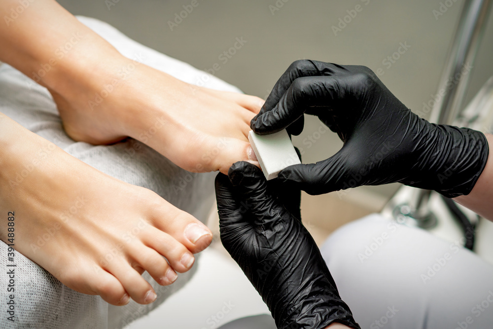 Hands of pedicurist buffing toenail of woman by white nail buff in nail salon