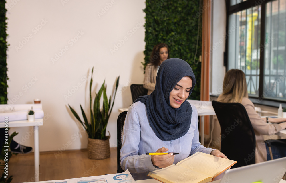 Portrait of a young muslim businesswoman working in an office while sitting at a table with colleagues in the background.