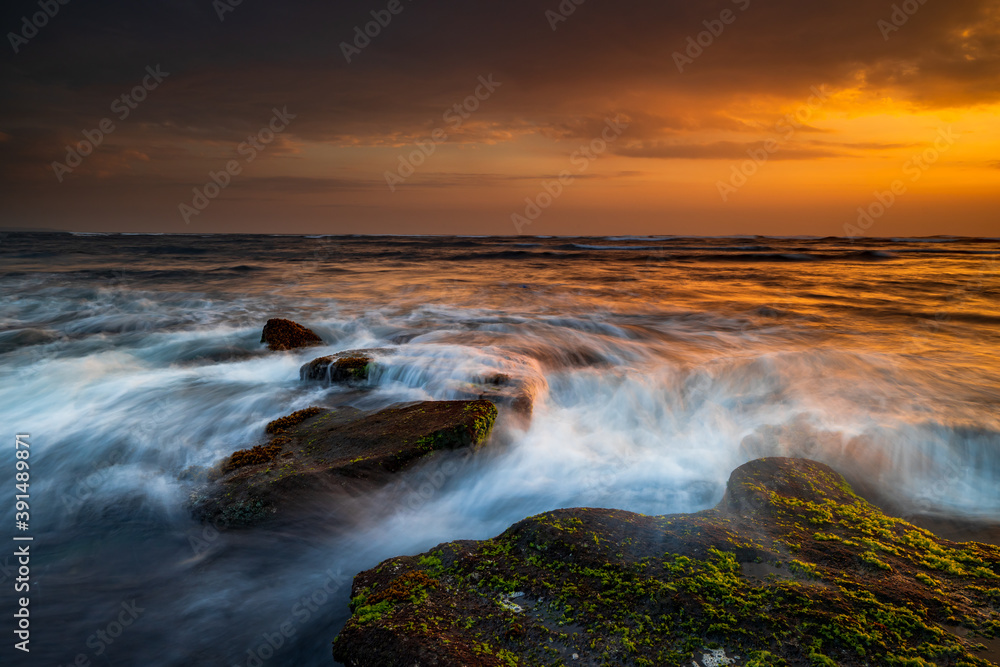 Obraz premium Beautiful seascape. Beach with stones covered by seaweeds. Low tide. Composition of nature. Motion water. Cloudy sky with sunlight. Slow shutter speed. Mengening beach, Bali, Indonesia