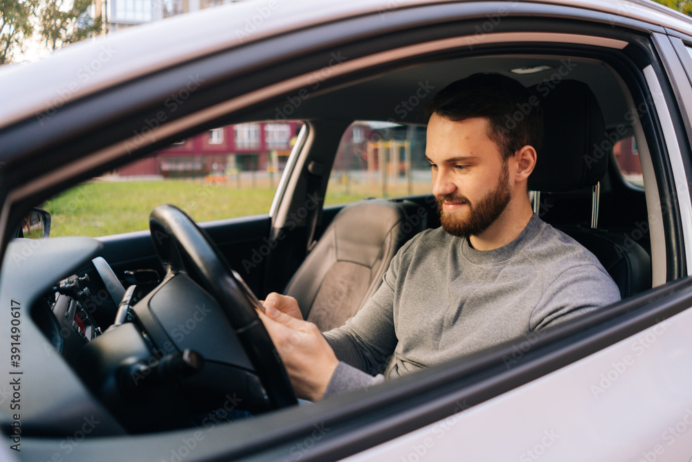 Caucasian bearded young man sitting in car and typing online message on cell phone, side view. Handsome businessman using mobile phone in auto. Male sitting in modern vehicle and works on smartphone.