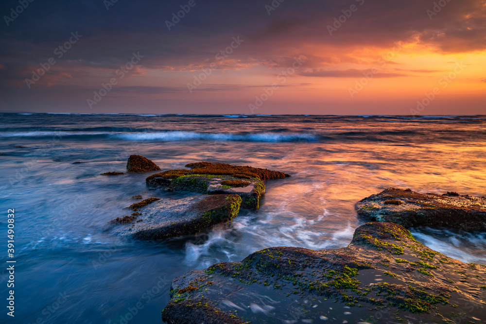 Obraz premium Beautiful seascape. Beach with stones covered by seaweeds. Low tide. Composition of nature. Motion water. Cloudy sky with sunlight. Slow shutter speed. Mengening beach, Bali, Indonesia