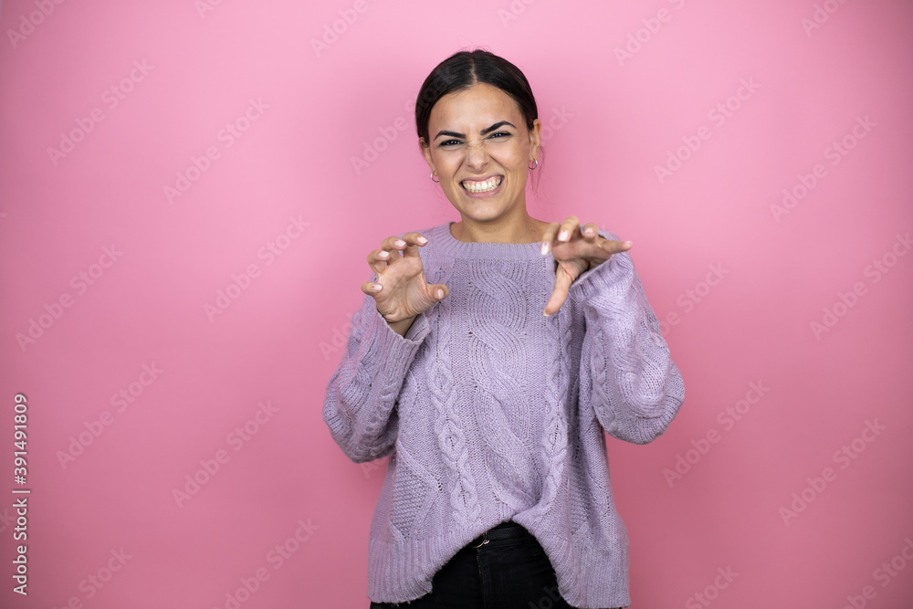 Beautiful woman wearing a casual violet sweater over pink background smiling funny doing claw gesture as cat, aggressive and sexy expression Beautiful woman wearing a casual violet sweater over pink background smiling funny doing claw gesture as cat, aggressive and sexy expression