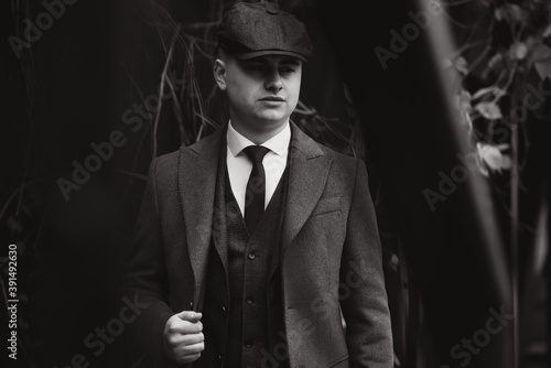 A man posing in the image of an English retro gangster of the 1920s dressed in a coat, suit and flat cap in Peaky blinders style.