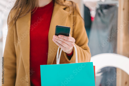 Cropped shot of stylish woman reading message on her smartphone while standing in a mall with a bunch of colorful paperbags in hands