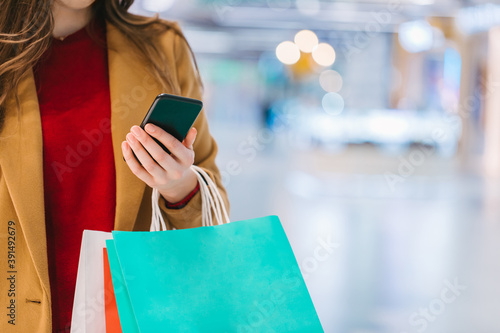 Cropped shot of elegant young woman using smartphone while standing in a mall with a bunch of colorful paperbags in hands