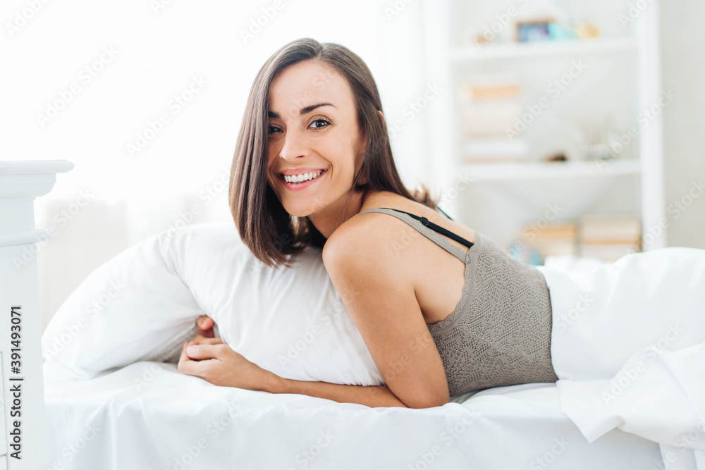 Smiling beautiful young woman lying in bed in modern light colored apartment in the morning Smiling beautiful young woman lying in bed in modern light colored apartment in the morning
