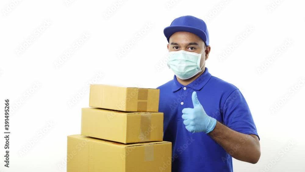 Young asian delivery man in blue uniform wearing protection mask and medical rubber gloves holding parcel cardboard and showing thumbs up on isolated. 4k Resolution. Young asian delivery man in blue uniform wearing protection mask and medical rubber gloves holding parcel cardboard and showing thumbs up on isolated. 4k Resolution.