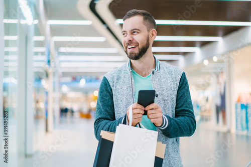 Handsome smiling young bearded man in casual outfit looking at the show-window while walking in a mall with a bunch of shopping bags and smartphone in hands