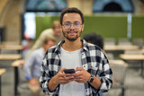 Portrait of a young happy mixed race man holding smartphone and looking at camera while working in the modern office, selective focus