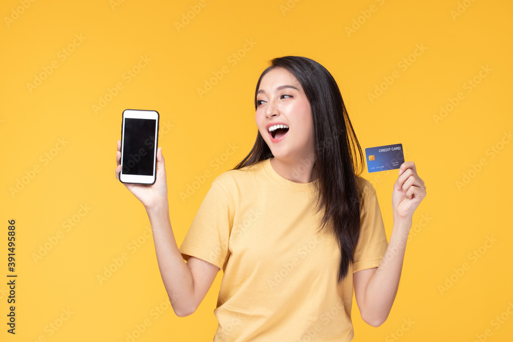 Portrait Asian happy young girl smiling cheerful and showing plastic credit card while holding mobile phone isolated on yellow background Portrait Asian happy young girl smiling cheerful and showing plastic credit card while holding mobile phone isolated on yellow background
