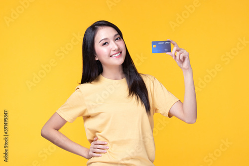 Cheerful young asian woman in yellow T-shirt holding bank card isolated on yellow background