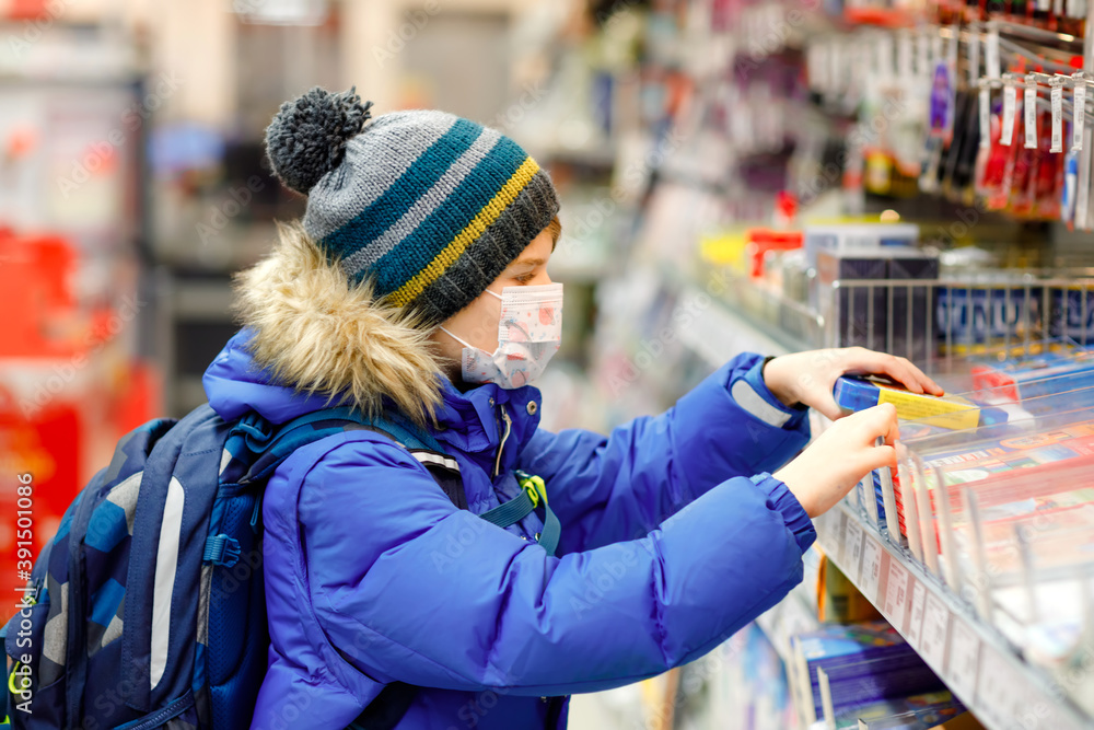 Little kid boy wearing medical mask buy school stuff in supermarket. Child with backpack and winter clothes. Schoolkid buying pencils. Lockdown and quarantine time during corona pandemic disease Little kid boy wearing medical mask buy school stuff in supermarket. Child with backpack and winter clothes. Schoolkid buying pencils. Lockdown and quarantine time during corona pandemic disease
