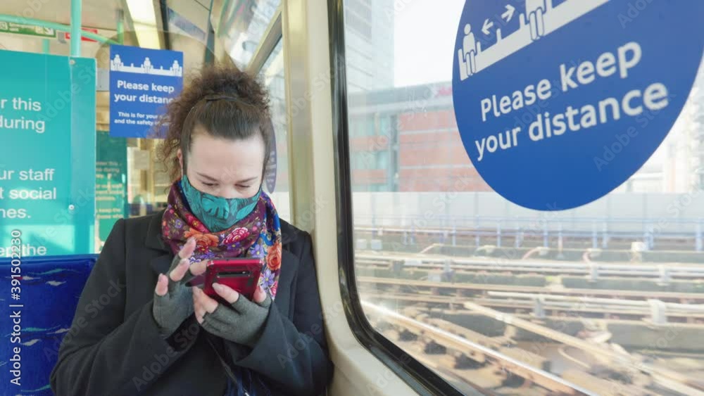 female passenger wearing face covering mask during covid-19 lockdown using phone inside train in england uk