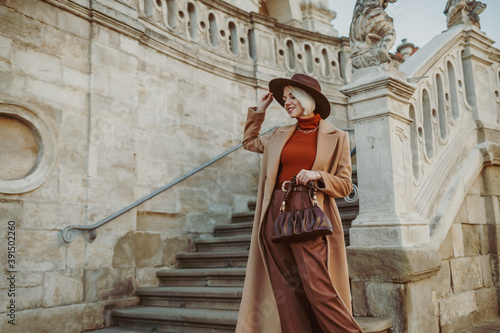 Outdoor autumn fashion portrait of elegant happy smiling woman wearing trendy midi beige, camel color coat, brown hat, trousers, orange turtleneck, holding leather pouch bag, posing in street of city © Victoria Fox