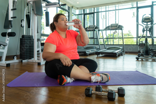 Asian fat woman fights overweight in the gym, doing heavy fitness exercises for future strong body. Obese person tired and drink water.