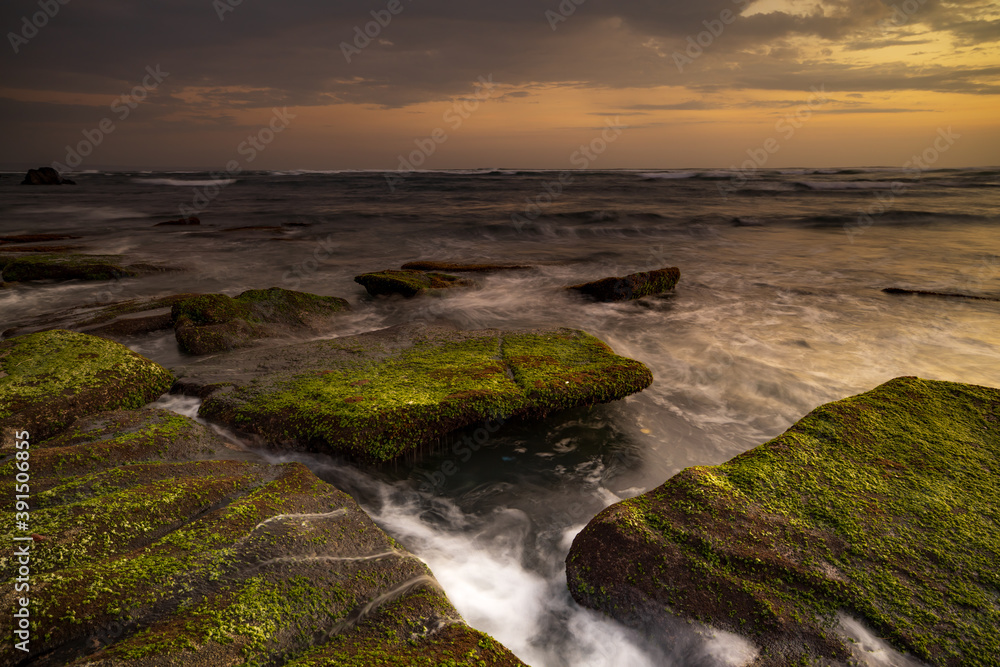 Obraz premium Calm ocean long exposure. Stones covered by green moss in mysterious mist of the sea waves. Concept of nature background. Sunset scenery background. Mengening beach, Bali, Indonesia.
