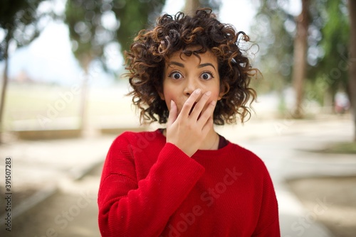 Photos Emotional Young arab woman wearing casual red sweater in the street gasps from astonishment, covers opened mouth with palm, looks shocked at camera