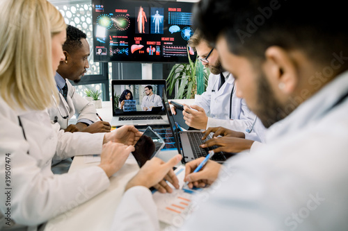 Team of five diverse multiethnic doctors having video conference meeting in hospital, chatting with their two young African and Caucasian colleagues, analyzing CT, ways of treatment and diagnostics.