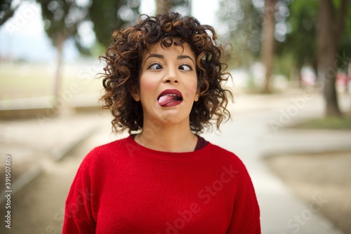 Young arab woman wearing casual red sweater in the street, showing grimace face crossing her eyes and showing tongue. Being funny and crazy