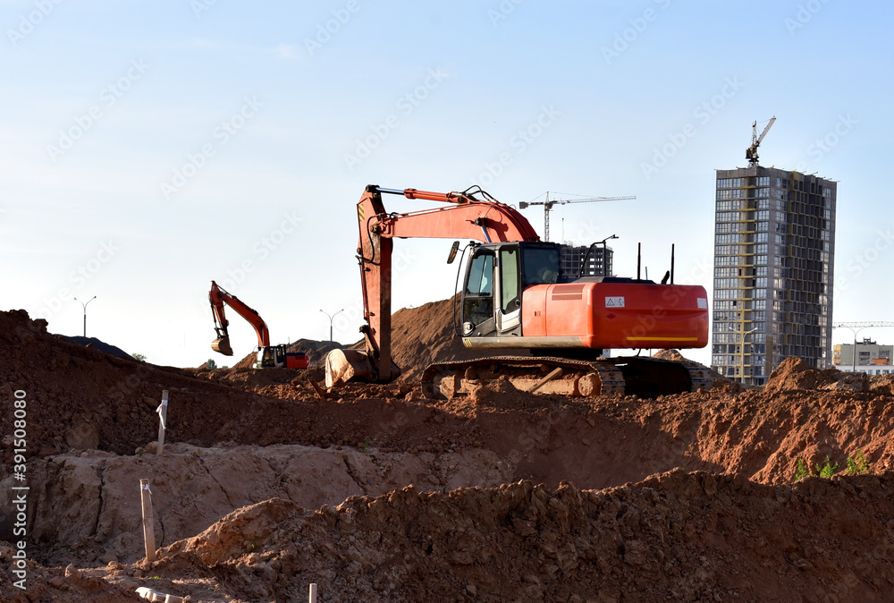 Excavator on earthworks at construction site. Backhoe on foundation work and road construction. Tower cranes in action on blue sky background. Heavy machinery and construction equipment Excavator on earthworks at construction site. Backhoe on foundation work and road construction. Tower cranes in action on blue sky background. Heavy machinery and construction equipment