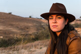 Young woman in hat and brown leather jacket, looking at camera, in natural park.